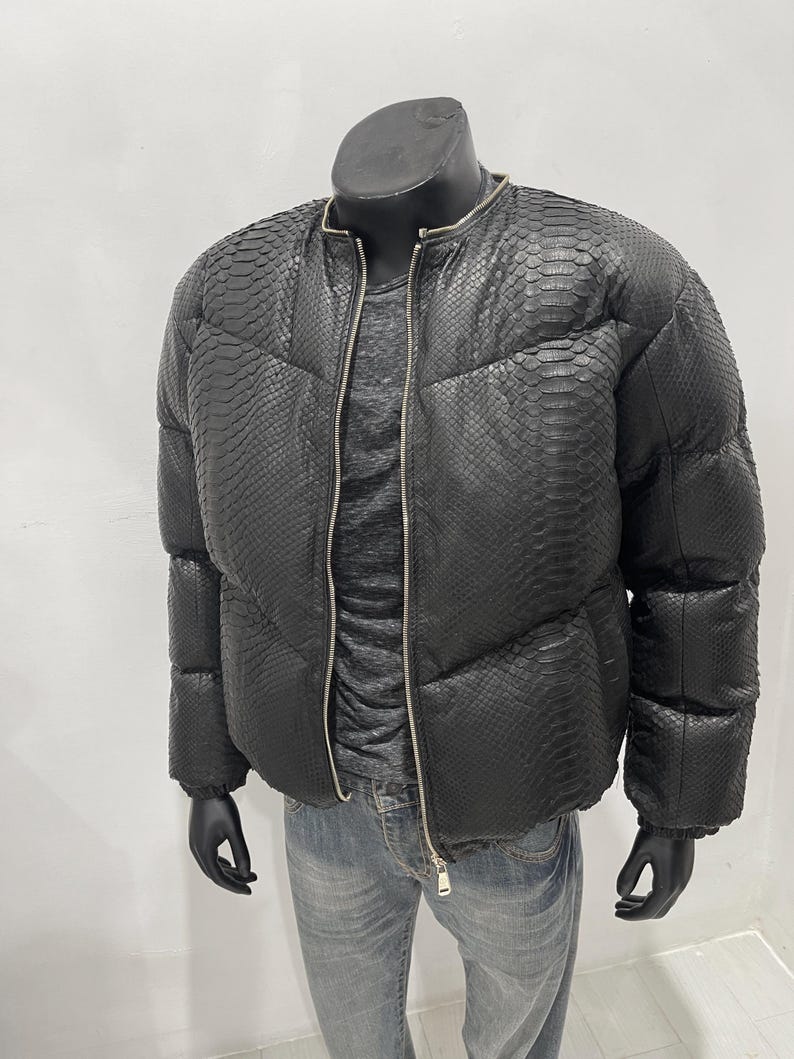Op de afbeelding: Zwarte pufferjack met slangenpatroon, ritssluiting en ronde kraag. Gepresenteerd op een mannequin, gecombineerd met een grijs shirt en een blauwe jeans. Het ontwerp van de jas omvat een gewatteerd patroon.