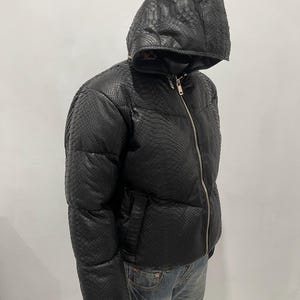 Op de afbeelding: Zwarte pufferjack met capuchon en slangenprint. De jas heeft een volledige ritssluiting en zakken. Het model draagt een blauwe jeans en zwarte handschoenen. Een stijlvol kledingstuk voor buiten.