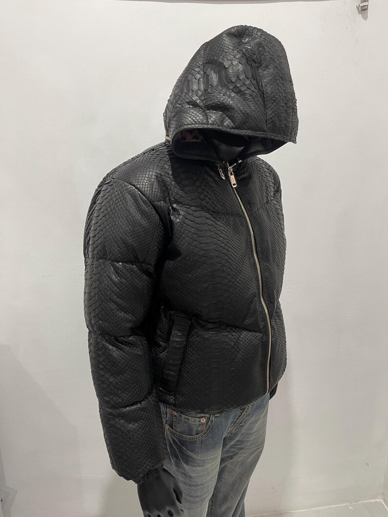 Op de afbeelding: Zwarte pufferjack met capuchon en slangenprint. De jas heeft een volledige ritssluiting en zakken. Het model draagt een blauwe jeans en zwarte handschoenen. Een stijlvol kledingstuk voor buiten.