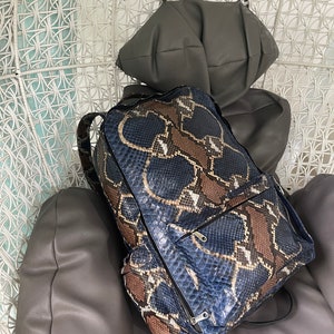 Op de afbeelding: Een blauwe en bruine slangleren rugzak met zwarte banden en ritsen. De rugzak staat op een grijze stoel.