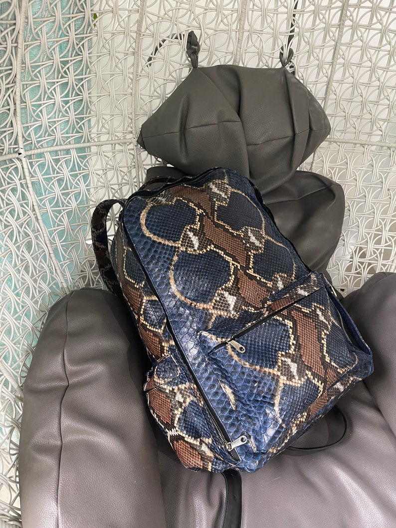 Op de afbeelding: Een blauwe en bruine slangleren rugzak met zwarte banden en ritsen. De rugzak staat op een grijze stoel.