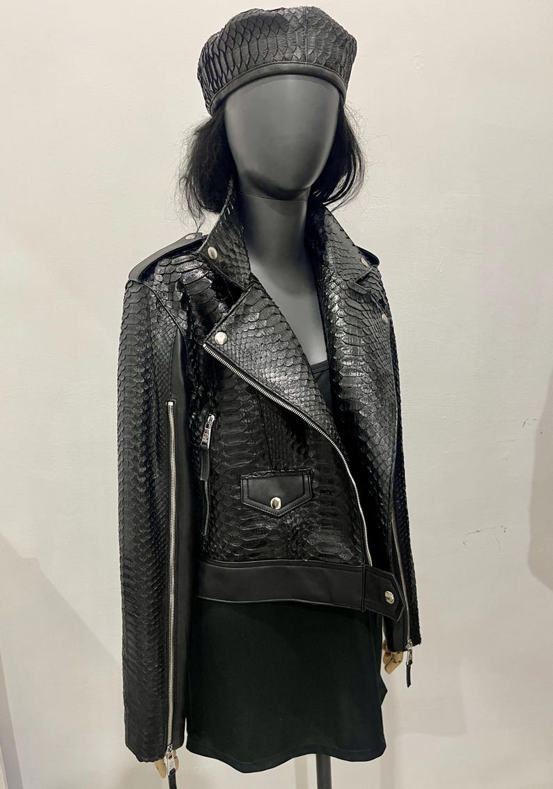 K&ouml;nnte beinhalten: Eine schwarze Lederjacke mit Schlangenhautmuster. Die Jacke hat einen Rei&szlig;verschluss, zwei Taschen und einen Kragen. Die Jacke wird von einer Puppe mit einem schwarzen Barett getragen.