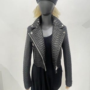 以下が含まれることがあります： ヘビ柄の黒い革のジャケット。ジャケットはジッパー留めと襟があります。ジャケットは黒いベレー帽をかぶったマネキンに着せられています。