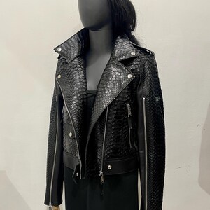 K&ouml;nnte beinhalten: Eine schwarze Lederjacke mit Schlangenhautmuster. Die Jacke hat einen Rei&szlig;verschluss und einen Stehkragen. Die Jacke ist kurz und hat einen Rei&szlig;verschlussdetail an den &Auml;rmeln.