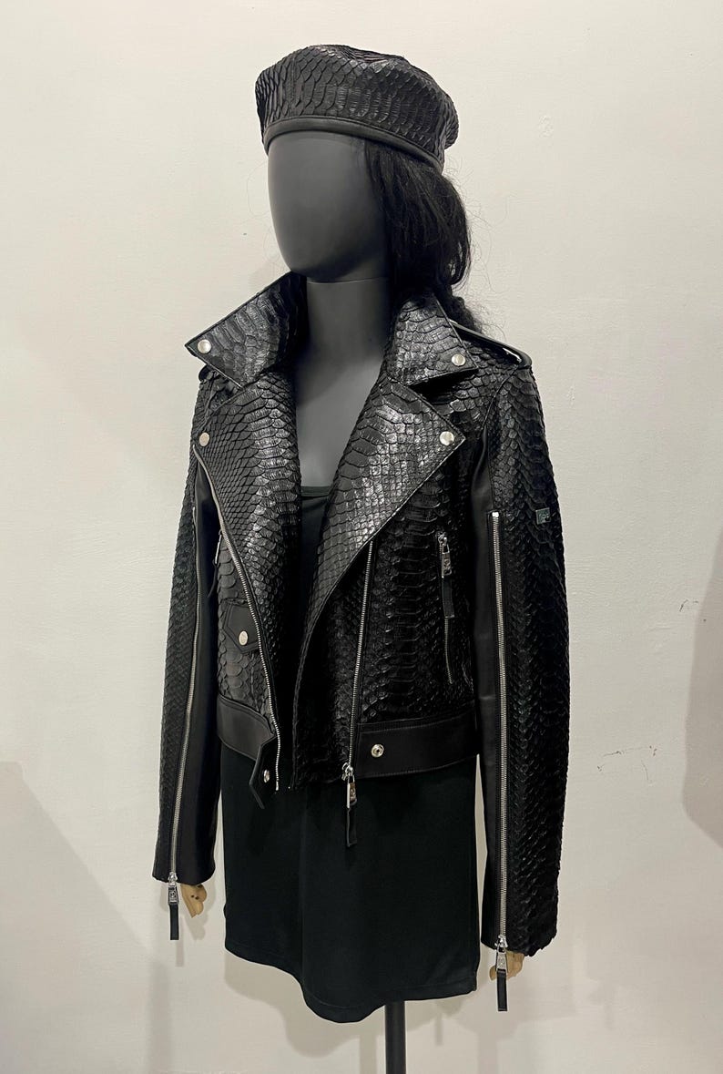 K&ouml;nnte beinhalten: Eine schwarze Lederjacke mit Schlangenhautmuster. Die Jacke hat einen Rei&szlig;verschluss und einen Stehkragen. Die Jacke ist kurz und hat einen Rei&szlig;verschlussdetail an den &Auml;rmeln.