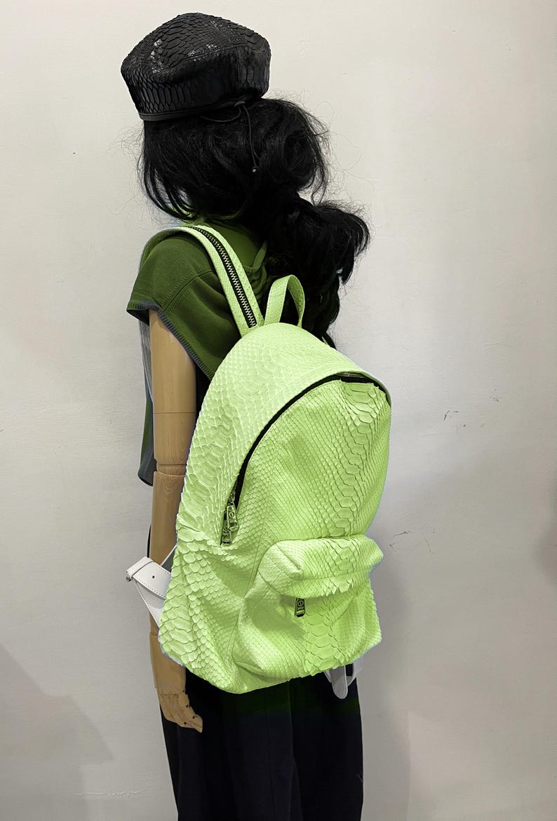 May include: A bright green snakeskin backpack with a zipper closure. The backpack is being worn by a mannequin with black hair and a black beret.
