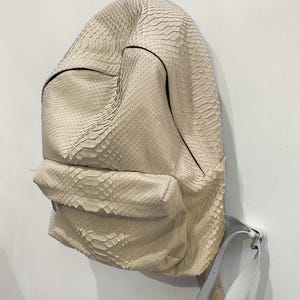 Op de afbeelding: Een lichtbeige rugzak van slangenleer met een witte leren band. De rugzak heeft een ritsvak aan de voorkant en een bovenste handvat.