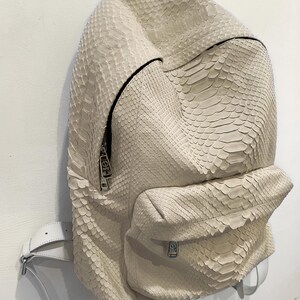 Op de afbeelding: Een witte slangenvellen rugzak met een ritsvak en een witte band.