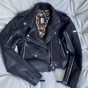 Może przedstawiać: Czarna sk&oacute;rzana kurtka motocyklowa z wzorem sk&oacute;ry węża i detalami z zamkiem błyskawicznym. Kurtka ma skr&oacute;coną długość i kołnierz.