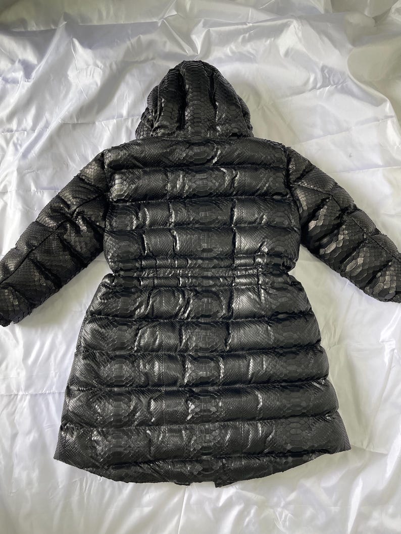 May include: Black puffer coat with a hood, featuring a snakeskin pattern. The coat has a cinched waist and a quilted design. The back view shows the full length of the coat, which is on a white background.