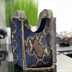 以下が含まれることがあります： 青、茶色、黒のヘビ柄のハンドバッグ。ゴールドのチェーンストラップとグレーのハンドルが付いています。バッグはバケツ型で、構造的な形状をしています。背景はぼやけていますが、キッチンのようです。