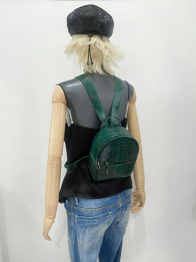 Peut inclure: Un sac &agrave; dos en cuir de serpent vert avec une fermeture &eacute;clair. Le sac &agrave; dos est pr&eacute;sent&eacute; sur un mannequin portant un haut noir et un jean bleu.