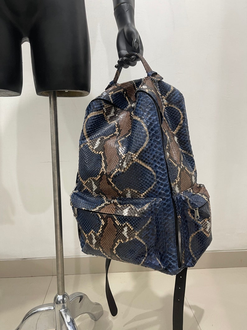 Op de afbeelding: Een blauwe en bruine slangleren rugzak met zwarte banden en een ritsvak aan de voorkant.