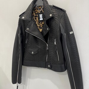 Może przedstawiać: Czarna sk&oacute;rzana kurtka motocyklowa z wzorem sk&oacute;ry węża. Kurtka ma zamek błyskawiczny, dwie kieszenie i detal z zamkiem błyskawicznym na rękawach.