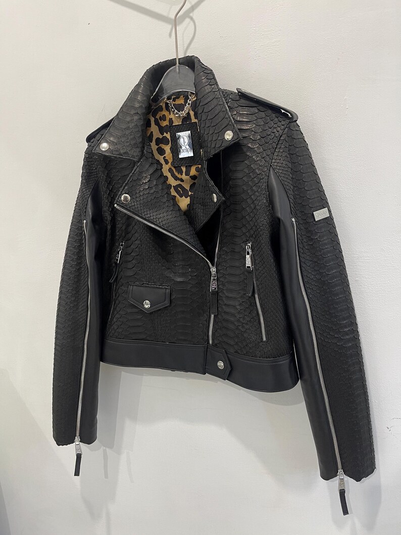 Może przedstawiać: Czarna sk&oacute;rzana kurtka motocyklowa z wzorem sk&oacute;ry węża. Kurtka ma zamek błyskawiczny, dwie kieszenie i detal z zamkiem błyskawicznym na rękawach.