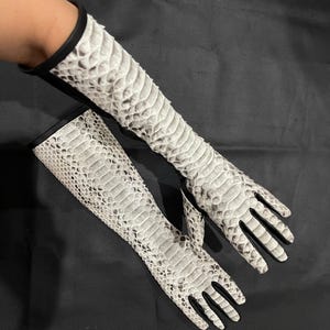 以下が含まれることがあります： 白とグレーのヘビ革模様のロンググローブのペア。手袋は手首に黒いトリムがあり、指なしです。手袋は爬虫類の革素材で作られており、黒い表面に表示されています。