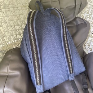K&ouml;nnte beinhalten: Ein blauer Rucksack aus Schlangenleder mit schwarzen Rei&szlig;verschl&uuml;ssen und verstellbaren Tr&auml;gern.