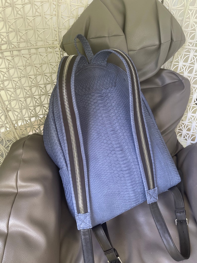 K&ouml;nnte beinhalten: Ein blauer Rucksack aus Schlangenleder mit schwarzen Rei&szlig;verschl&uuml;ssen und verstellbaren Tr&auml;gern.