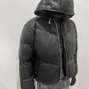 Op de afbeelding: Zwarte pufferjack met capuchon, met een slangenleerpatroon. De jas heeft een ritssluiting en wordt op een mannequin getoond. De mannequin draagt ook een blauwe spijkerbroek.