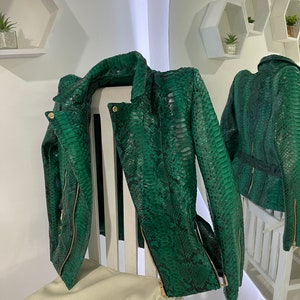 May include: A green snakeskin leather jacket with gold zippers. The jacket is hanging on a white chair with a white bag on the seat.