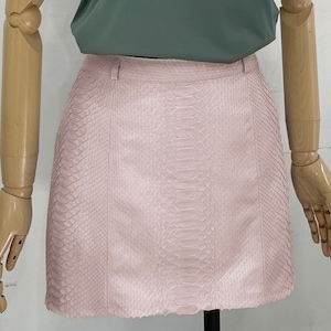 Peut inclure: Une mini-jupe en cuir de serpent rose avec une fermeture éclair. La jupe est en cuir véritable et a un léger brillant.