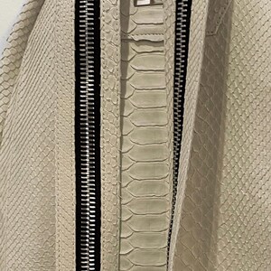 Op de afbeelding: Een witte slangleren rugzak met twee zwarte ritsen. De rugzak heeft een klein label met het woord "Belp" erop.