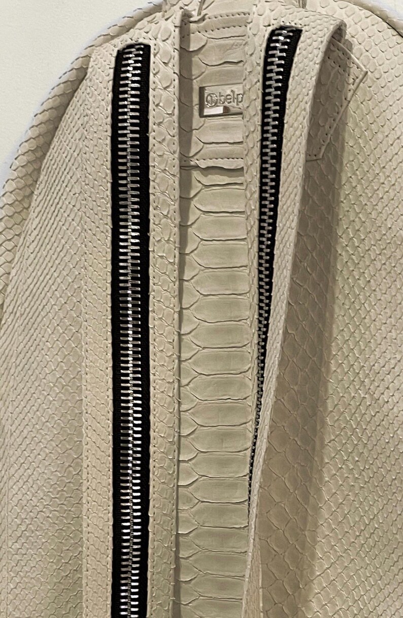 Op de afbeelding: Een witte slangleren rugzak met twee zwarte ritsen. De rugzak heeft een klein label met het woord "Belp" erop.