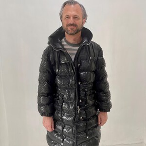 May include: A long, black puffer coat with a snakeskin pattern. The coat has a hood, a zipper closure, and pockets. The person in the image is wearing a striped shirt underneath the coat.