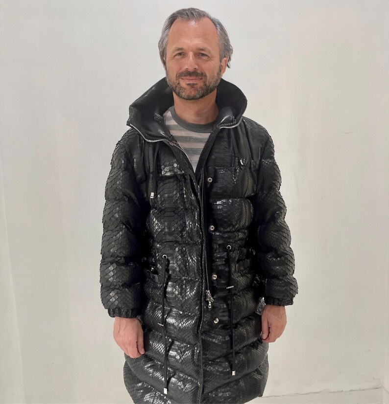 May include: A long, black puffer coat with a snakeskin pattern. The coat has a hood, a zipper closure, and pockets. The person in the image is wearing a striped shirt underneath the coat.