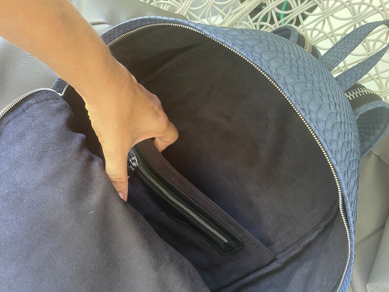 K&ouml;nnte beinhalten: Ein blauer Lederrucksack mit einer Rei&szlig;verschlusstasche auf der Vorderseite. Der Rucksack ist offen und das Innere ist mit einem dunklen Stoff ausgekleidet.