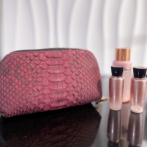 Peut inclure: Une trousse de maquillage en cuir de serpent de couleur bordeaux avec une fermeture éclair, à côté de deux petits flacons de parfum avec des bouchons dorés et du verre transparent.