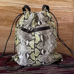 以下が含まれることがあります： 黒のストラップが付いた、薄い緑色と灰色のヘビ革製の巾着バックパックです。