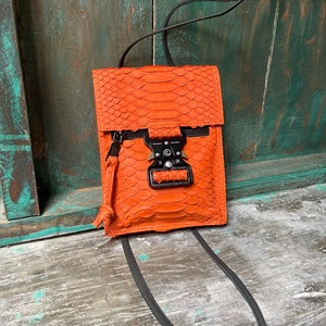 Peut inclure: Un petit sac bandoulière en peau de serpent orange avec une bandoulière noire et une fermeture à boucle argentée.
