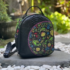 May include: Black backpack with a floral design. The backpack has a textured, snake-like pattern and a top handle. The floral design features green, yellow, blue, and pink. The backpack has adjustable straps.