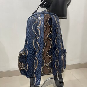 Op de afbeelding: Een blauwe en bruine slangleren rugzak met verstelbare banden. De rugzak heeft een ritsvak aan de voorkant en een ritsvak aan elke zijde.