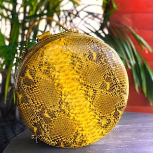 May include: A circular, yellow handbag featuring a snakeskin design. The bag has a zip closure and a small handle. The bag is approximately 20 cm in diameter.