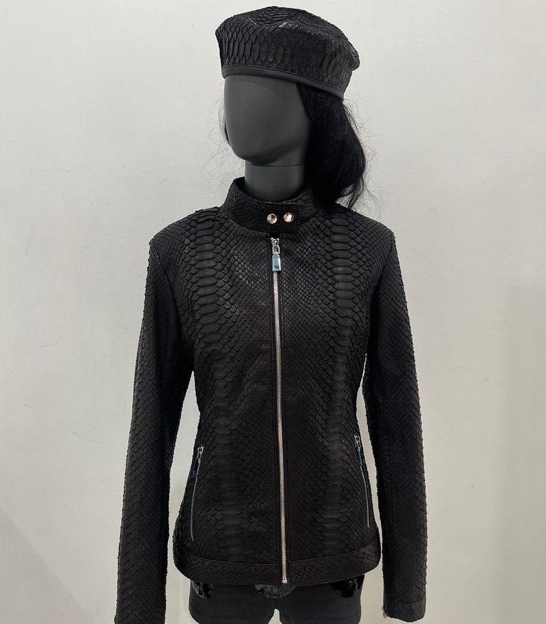 May include: A black leather jacket with a snakeskin pattern. The jacket has a zip fastening and two pockets on the sides. The jacket is being modelled on a mannequin wearing a black beret.