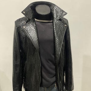 May include: Black faux leather jacket with a snakeskin texture, featuring a notched collar, zipper closure, and a belt at the waist. The jacket is displayed on a black mannequin, paired with a black shirt and blue jeans.