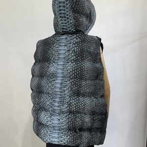 May include: A black and grey puffer vest with a snakeskin pattern. The vest has a hood and is sleeveless.