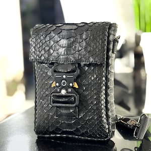 May include: A black leather crossbody bag with a snakeskin pattern and a gold buckle closure. The bag has a black adjustable strap.