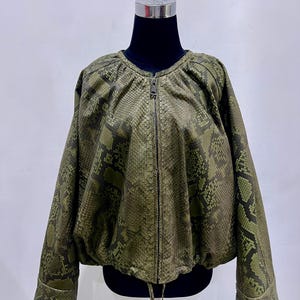 May include: Olive green jacket with a snakeskin pattern. The jacket has a zip-up front, long sleeves, and a drawstring at the waist. The jacket is displayed on a black mannequin.