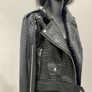 K&ouml;nnte beinhalten: Schwarze Lederjacke mit Schlangenhautmuster. Die Jacke hat einen Rei&szlig;verschluss, einen Kragen und zwei Taschen.