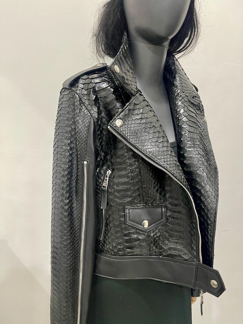 K&ouml;nnte beinhalten: Schwarze Lederjacke mit Schlangenhautmuster. Die Jacke hat einen Rei&szlig;verschluss, einen Kragen und zwei Taschen.