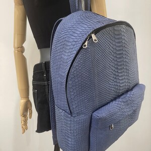 K&ouml;nnte beinhalten: Ein blauer Schlangenleder-Rucksack mit einer Rei&szlig;verschlusstasche vorne und einem silbernen Rei&szlig;verschluss-Zipper. Der Rucksack hat eine strukturierte, schuppige Oberfl&auml;che.