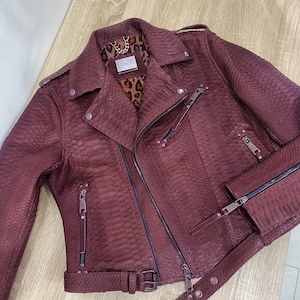 May include: A burgundy leather motorcycle jacket with a zipper closure and a snakeskin pattern. The jacket has a belt at the waist and a zippered pocket on the left sleeve. The label on the inside of the jacket reads "Dielly".