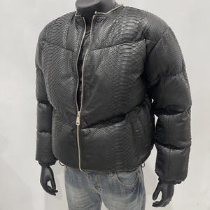 Op de afbeelding: Zwarte pufferjack met een slangenpatroon, met een zilveren ritssluiting. De jas wordt getoond op een mannequin met zwarte handschoenen en gecombineerd met een blauwe jeans. De jas heeft een ronde kraag.