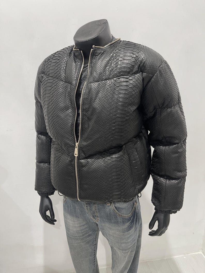 Op de afbeelding: Zwarte pufferjack met een slangenpatroon, met een zilveren ritssluiting. De jas wordt getoond op een mannequin met zwarte handschoenen en gecombineerd met een blauwe jeans. De jas heeft een ronde kraag.