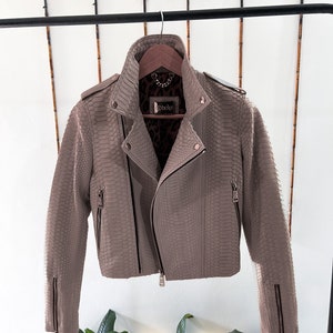 以下が含まれることがあります： ヘビ柄の薄い茶色の革のジャケット。ジャケットはジッパー留めと襟があります。ジャケットはハンガーにかけられています。