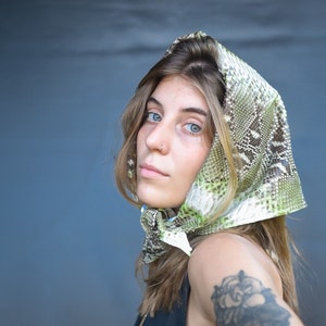 May include: A woman with long brown hair is wearing a green and brown patterned headscarf. The headscarf is made of a snakeskin-like material. The woman is looking at the camera.