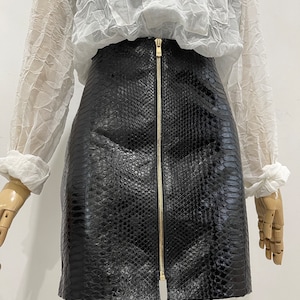 May include: A black faux snakeskin skirt with a gold zip. The skirt is paired with a white, sheer, long-sleeved blouse. The skirt has a textured pattern and a fitted silhouette.